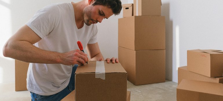 Young man using a red marker to label a cardboard box while preparing for a move surrounded by other packed boxes.