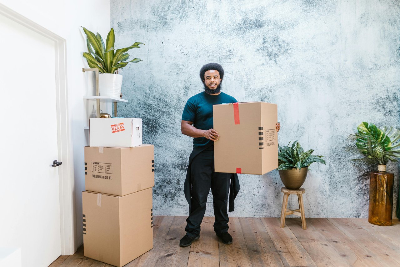 Man holding a moving box while standing beside stacked boxes and indoor plants.