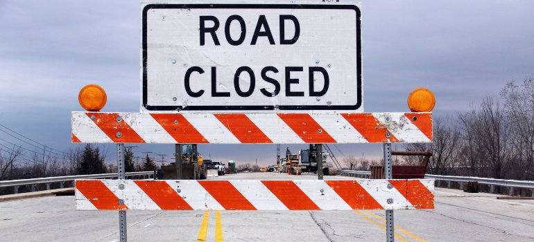 Orange and white striped road closure sign blocking off a street with construction vehicles visible in the background.