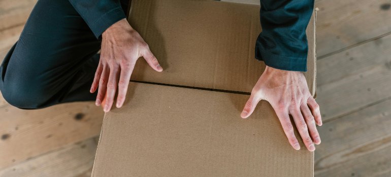 Close-up of a person’s hands sealing a cardboard moving box on a wooden floor.