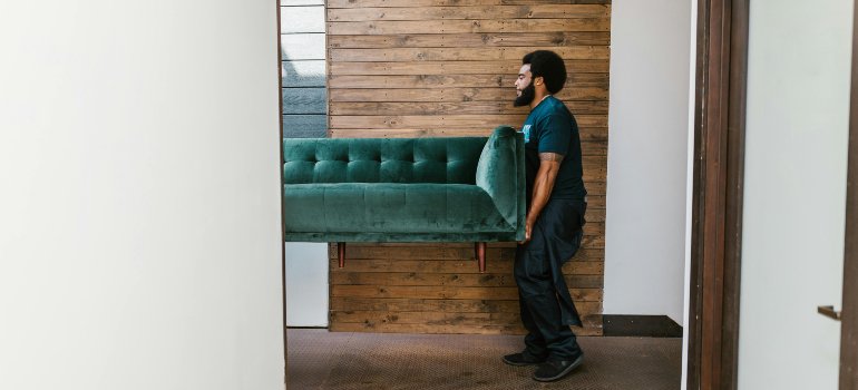 Professional mover lifting a green velvet sofa through a narrow hallway with wooden panel walls.