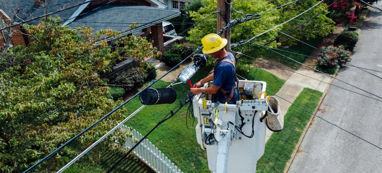 Utility worker in a lift repairing electrical lines above a suburban neighborhood with trees and houses below.