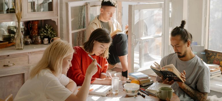 Four young adults sit in a cozy, sunlit apartment, reading and studying together at a cluttered table filled with books, mugs, and fruit.