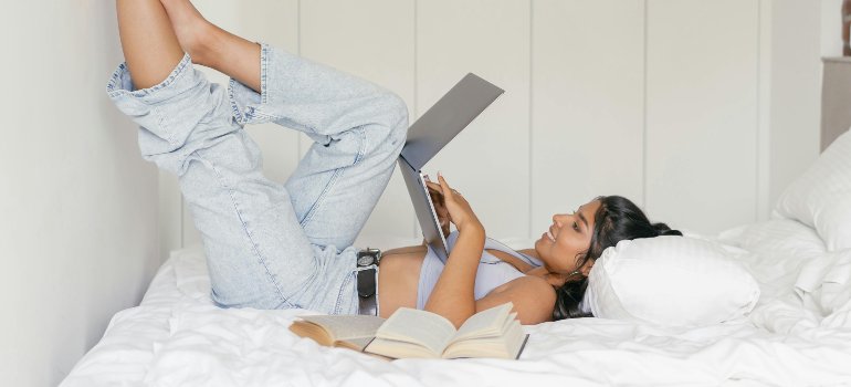 Young woman relaxing on her bed with a laptop and open books, enjoying her personal space after leaving her parents’ home for the first time.