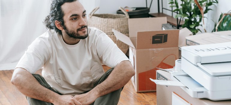 Young man sitting on the floor surrounded by moving boxes, reflecting on the changes after leaving his parents’ home for the first time.