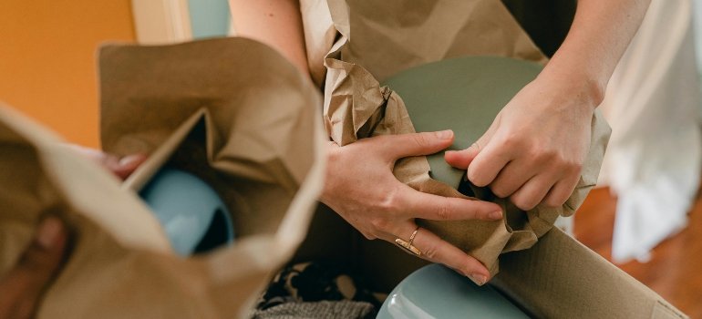 Close-up of hands wrapping ceramic dishes in brown paper for a careful pack.