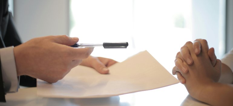 Person offering a pen for someone to sign paperwork across a table.
