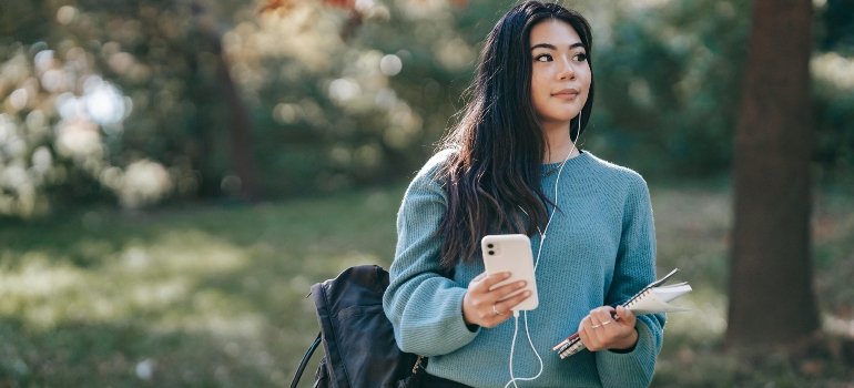 Asian woman in earphones with notebook using smartphone in park