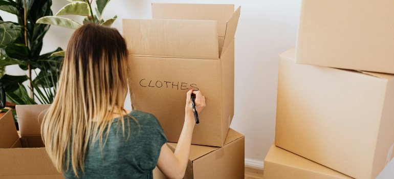 Woman writing 'clothes' on a cardboard moving box while packing at home.