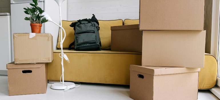 Moving boxes, lamp, and backpack placed near a yellow sofa in a modern apartment.