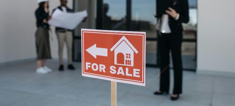 Red for sale sign in front of a property with people discussing in the background.