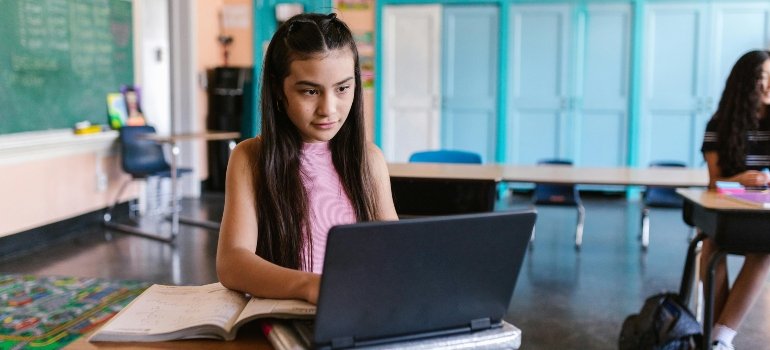 A Young Girl Typing on Her Laptop while Sitting on the Chair Inside the Classroom