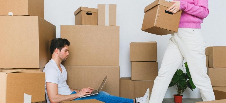 A woman is arranging boxes during relocation while a man is sitting on the floor searching for budget-friendly moving from Shavano Park to Floresville on his laptop.
