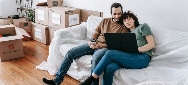 A couple sitting on a couch beside packed boxes, using a laptop to search for reliable movers to plan their upcoming relocation.