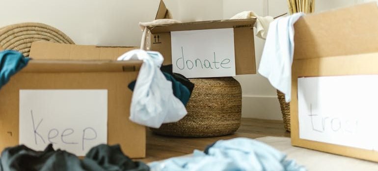 Three labeled cardboard boxes in a tidy room: 'keep,' 'donate,' and 'trash,' surrounded by discarded clothing items.