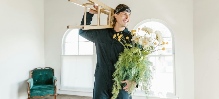 Man in a Jumpsuit Wearing a Bandana Carrying a Wooden Stool and Flowers in a Vase