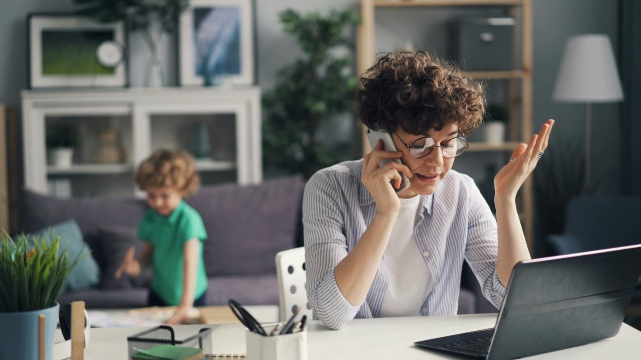 Woman Working on Laptop and Talking on Phone and Son Playing behind